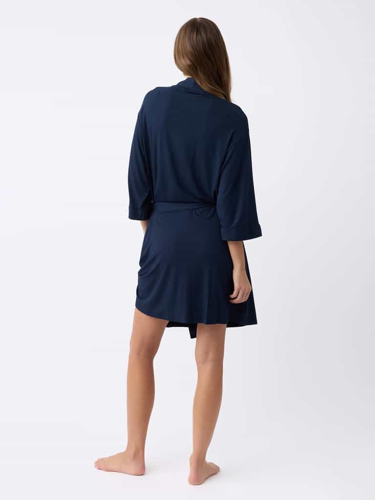 A person with long hair stands barefoot on a plain white background, facing away while wearing Cozy Earth's Women's Stretch-Knit Bamboo Kimono Robe in dark blue with three-quarter sleeves. |Color:Navy