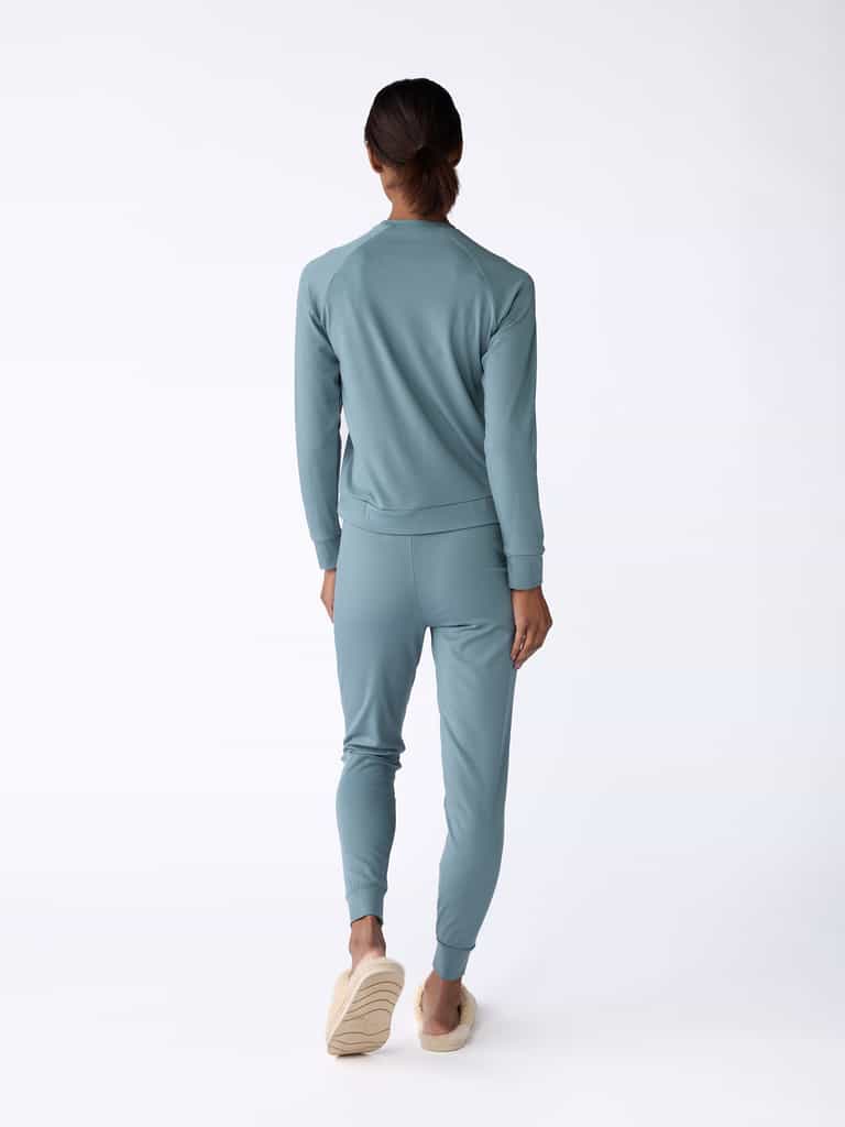 A woman is seen facing away, dressed in a matching blue-green tracksuit, featuring the Women's Bamboo Jogger Pant by Cozy Earth, complemented by beige slip-on shoes. The plain white background highlights the clothing ensemble. |Color:Oceanside