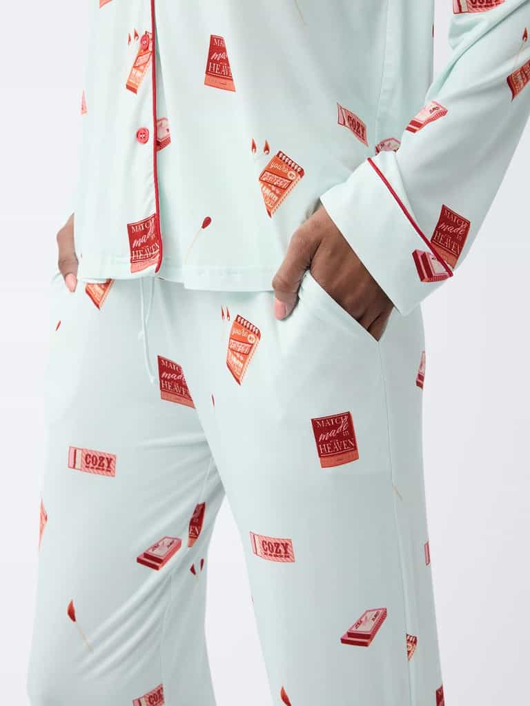 A person wearing the Last Chance Women's Bamboo Stretch-Knit Long Sleeve Pajama Set by Cozy Earth stands against a plain background. The pajamas are light blue with red matchbox patterns, featuring red buttons and piping. The person's left hand rests in the pants pocket. |Color:Perfect Match
