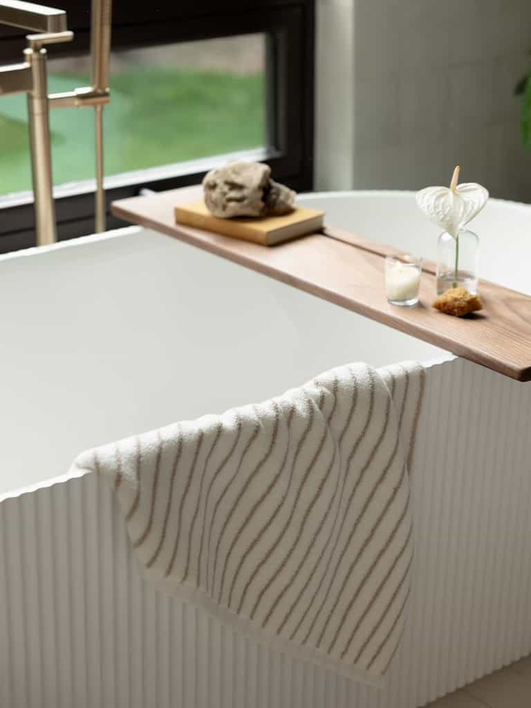 A modern white bathtub with a textured exterior stands near a window. A wooden tray spans the tub, holding two glass vases with plants, a candle, and a decorative stone. Draped over the tub's edge is a Luxe Bath Towel featuring thin, wavy brown stripes from Cozy Earth. |Color:Sand Stripe