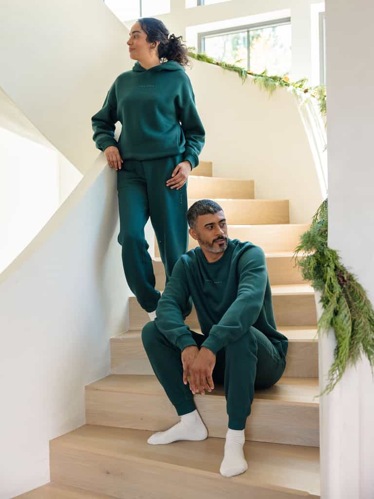 Two individuals, both donning the Men's CityScape Jogger by Cozy Earth in matching green, unwind on wooden stairs decorated with lush greenery. One person is seated on a step while the other stands with one foot on the step, gazing into the distance in a sunlit room. |Color:Spruce