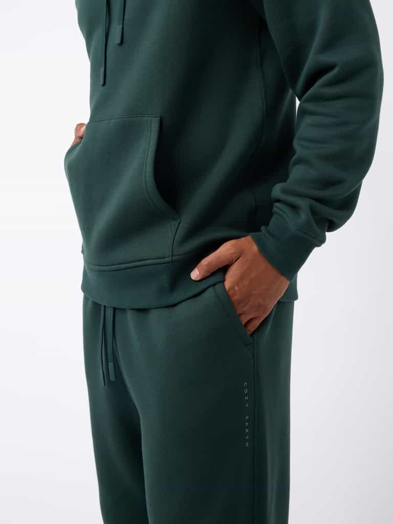 A person is wearing a dark green hoodie and matching Men's CityScape Sweatpants from Cozy Earth, with hands in pockets. The clothing features a subtle logo that reads "COZY EARTH" on the pants, set against a plain white background. |Color:Spruce