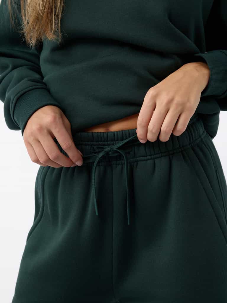 Close-up of a person adjusting the drawstring on Cozy Earth's Women's CityScape Wide Leg Pant in dark green. The individual is wearing a matching dark green sweatshirt against a plain white background. |Color:Spruce
