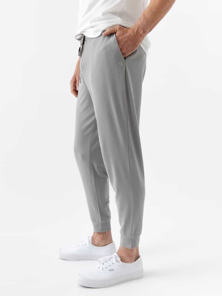 A person is wearing Cozy Earth's Men's Bamboo Jogger Set in gray with a white T-shirt, standing with one hand in their pocket and sporting white sneakers against a plain white background. |Color:Stone