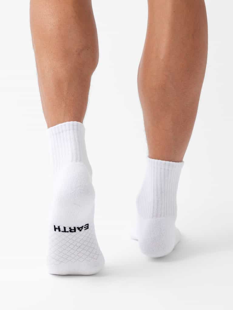 A person wearing Cozy Earth Essential Quarter Socks stands on a white background, back of legs visible. The word "EARTH" is printed in black near the heel on the sole of the left sock. |Color:White