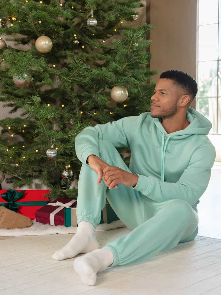 A man wearing a Cozy Earth's Men's CityScape hoodie in teal sits on the floor by a Christmas tree adorned with silver and gold ornaments. Several wrapped gifts are placed under the tree, as soft light filters through a nearby window. |Color:Wintergreen