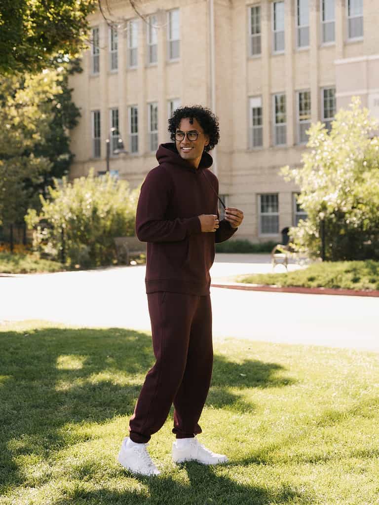 A person with curly hair, wearing glasses, and dressed in a maroon Men's CityScape Hoodie and matching sweatpants from Cozy Earth, stands smiling on a green lawn in front of a beige brick building. Bright sunlight casts shadows, and trees and bushes are visible in the background. |Color:Burgundy