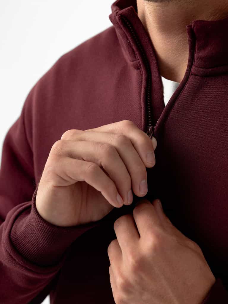 Close-up of a person zipping up a Cozy Earth Men's CityScape Quarter Zip sweater. The person's hands are visible, one holding the zipper pull while the other steadies the maroon fabric. The background is plain white. |Color:Burgundy