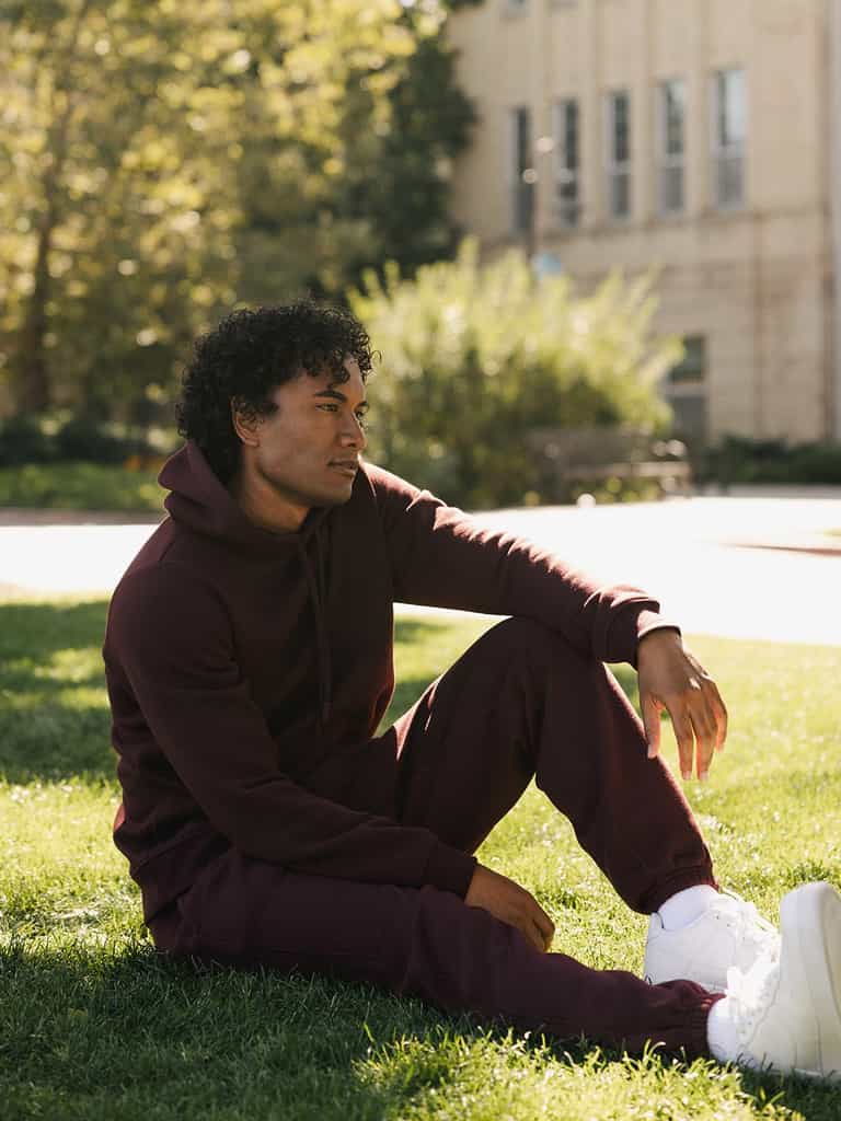 A person with curly hair is sitting on grassy ground in a relaxed pose, wearing a maroon hoodie, Men's CityScape Sweatpant by Cozy Earth, and white sneakers. The background features a tree-lined pathway and a building with large windows, bathed in sunlight. |Color:Burgundy