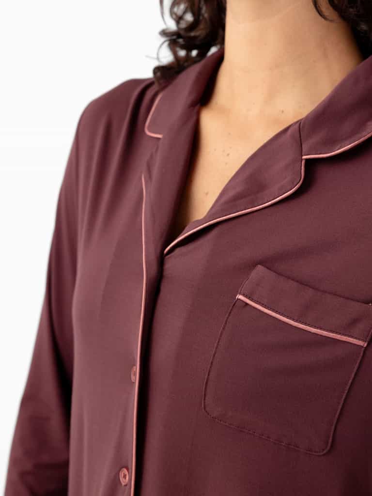 A close-up shows a person in Cozy Earth's Women's Bamboo Stretch-Knit Long Sleeve Pajama Set, featuring maroon fabric with pink piping, a chest pocket, and long sleeves. The person's head is partially out of the frame. |Color:Burgundy