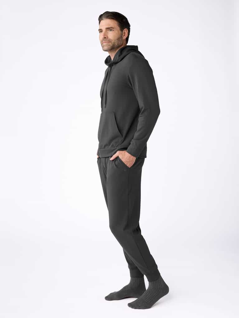 A man with short brown hair and a beard stands sideways against a white background, wearing the Cozy Earth Men's Brushed Bamboo Hoodie & Jogger Set in dark gray with gray socks. |Color:Charcoal