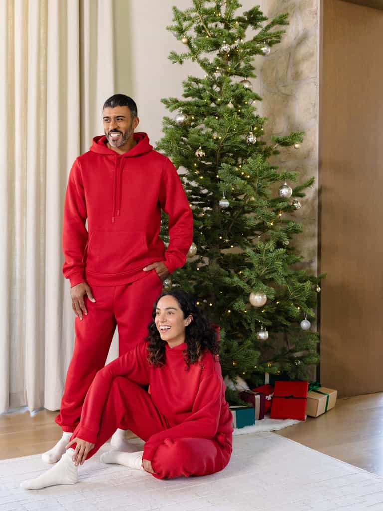 A man and a woman wearing matching Men's CityScape Hoodies from Cozy Earth are in a cozy living room decorated for Christmas. The woman sits on the floor, smiling, while the man stands behind her. A Christmas tree adorned with ornaments and presents is visible in the background. |Color:Crimson