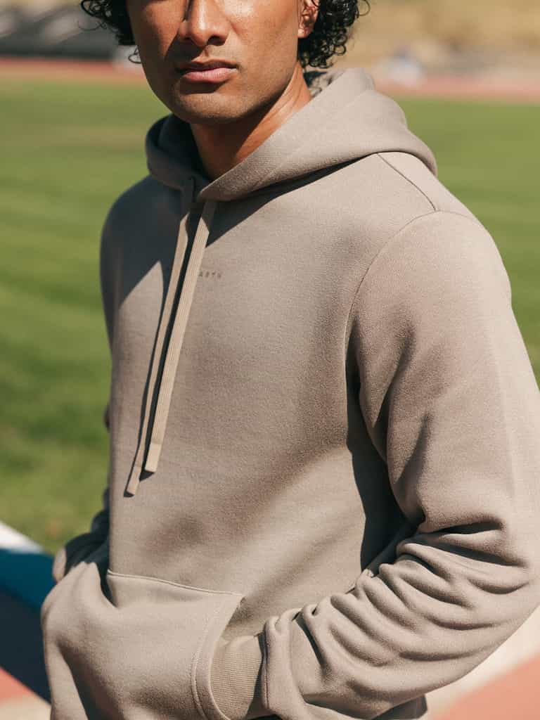 A person with curly hair stands outside on a sunny day wearing a beige Men's CityScape Hoodie by Cozy Earth, with their hands in the front pockets. The background is a blurred field and walking track. |Color:Ebony