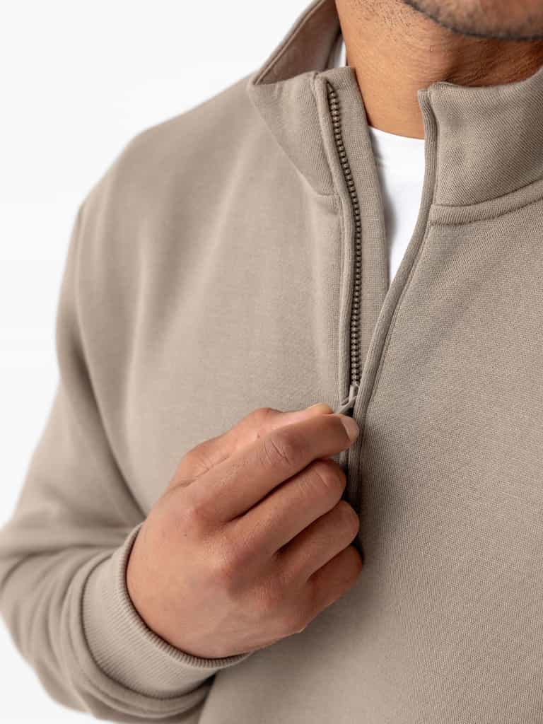 A person zips up a Cozy Earth Men's CityScape Quarter Zip in beige, revealing a glimpse of a white shirt underneath. The close-up focuses on the hand and the zipper, highlighting the texture and color of the sweater. The background is plain white. |Color:Ebony