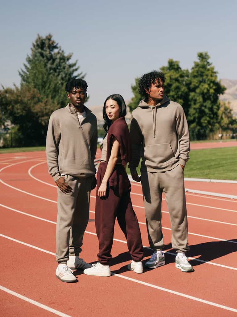Three people stand on an outdoor track under a clear sky. Two individuals wear beige Men's CityScape Sweatpants from Cozy Earth, while the person in the middle wears maroon Men's CityScape Sweatpants from Cozy Earth. They all wear white sneakers and pose confidently with trees and a distant hill in the background. |Color:Ebony