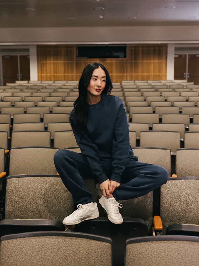 A person with long dark hair is sitting casually on the backrest of a seat in an empty auditorium. They are wearing a matching navy blue Women's CityScape Crewneck from Cozy Earth and white sneakers, with both feet resting on the seat in front of them. The setting is spacious and well-lit. |Color:Eclipse