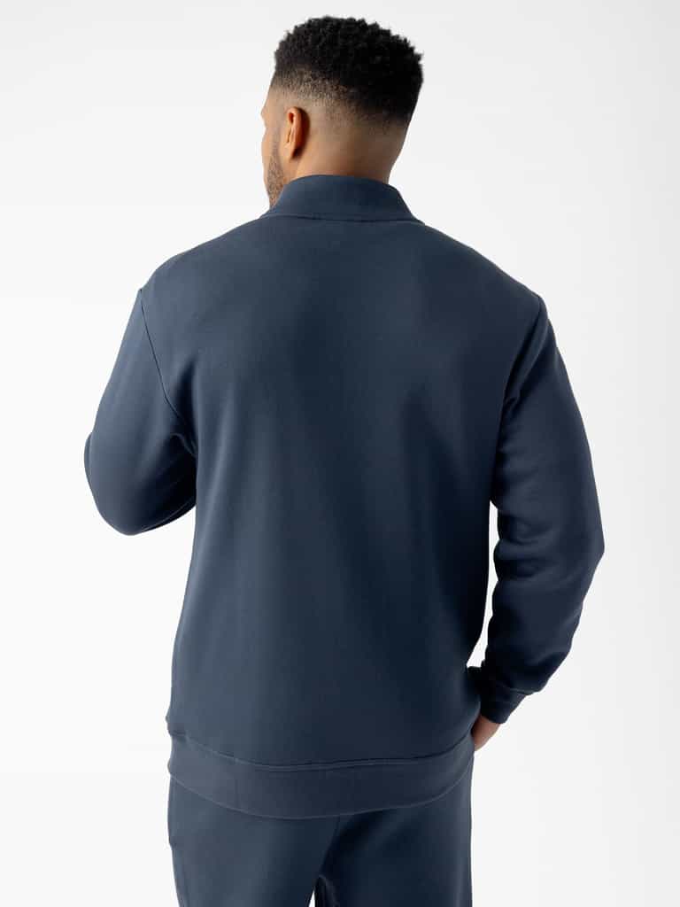 A person with short, curly hair is standing facing away from the camera, wearing a dark blue Men's CityScape Quarter Zip and matching pants from Cozy Earth. The background is plain white. |Color:Eclipse