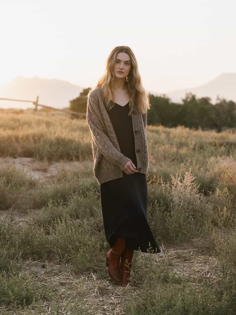 A woman with long, wavy hair stands in an open field at sunset. She is wearing Cozy Earth's Women's Sunset Slip Dress paired with a chunky knit cardigan and brown boots. A wooden fence and trees are visible in the background, along with distant mountains. |Color:Eclipse