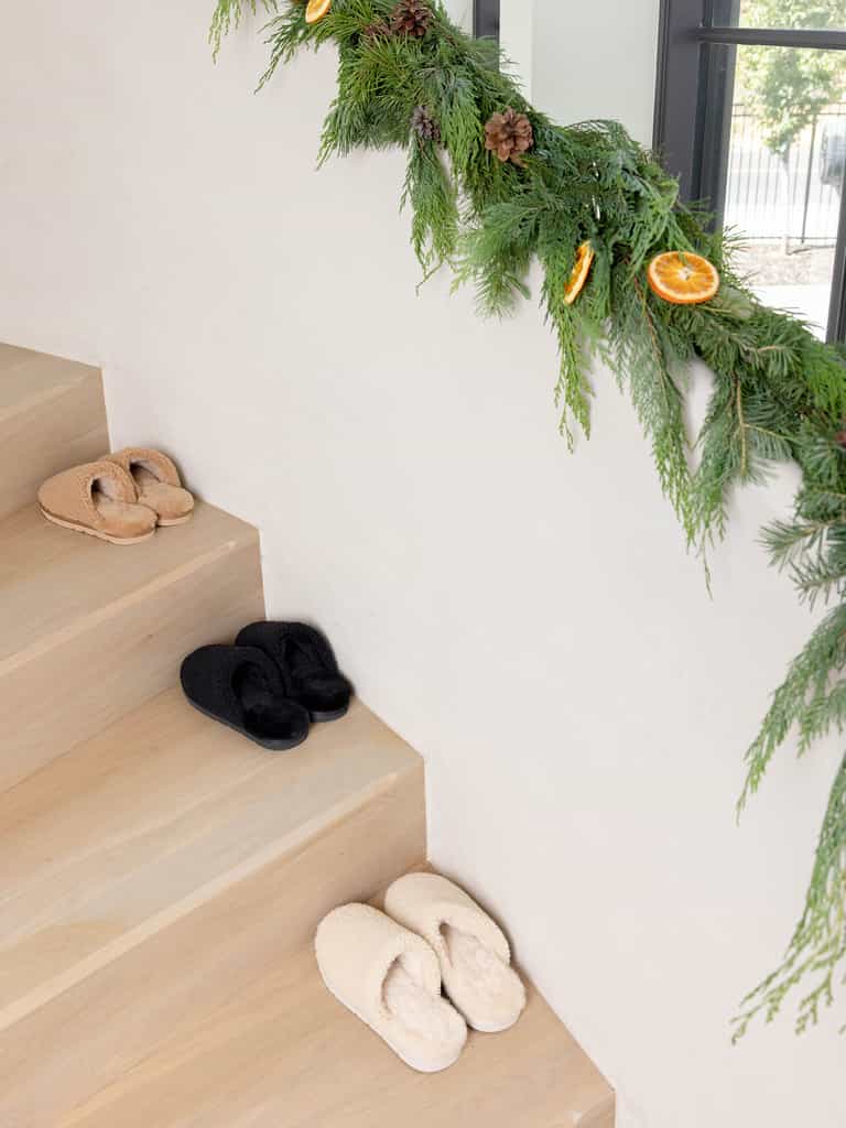 Three pairs of "Puffy Sheep Slipper" by Cozy Earth in beige and black are placed on wooden stairs. The railing is adorned with a garland of greenery featuring pinecones and dried orange slices, while bright light filters through a nearby window. |Color:Fawn