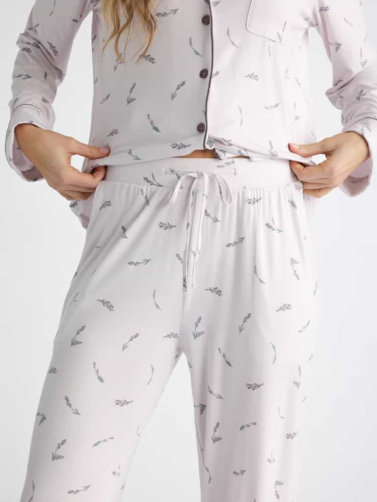 A person adjusts the waistband of Cozy Earth Women's Bamboo Stretch-Knit Pajama Pant, featuring a light color with small floral patterns. The top boasts buttons, a pocket, and both pieces offer a loose, comfortable fit. |Color:French Lavender