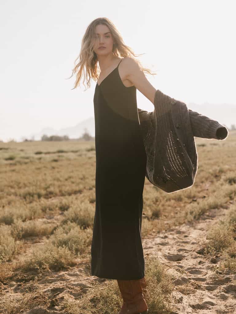 A woman with long wavy hair stands in a field, wearing the Women's Sunset Slip Dress by Cozy Earth and holding a gray knit cardigan. She gazes into the distance with a serene expression as the sun backlights her, casting a warm glow over the scene. |Color:Jet Black
