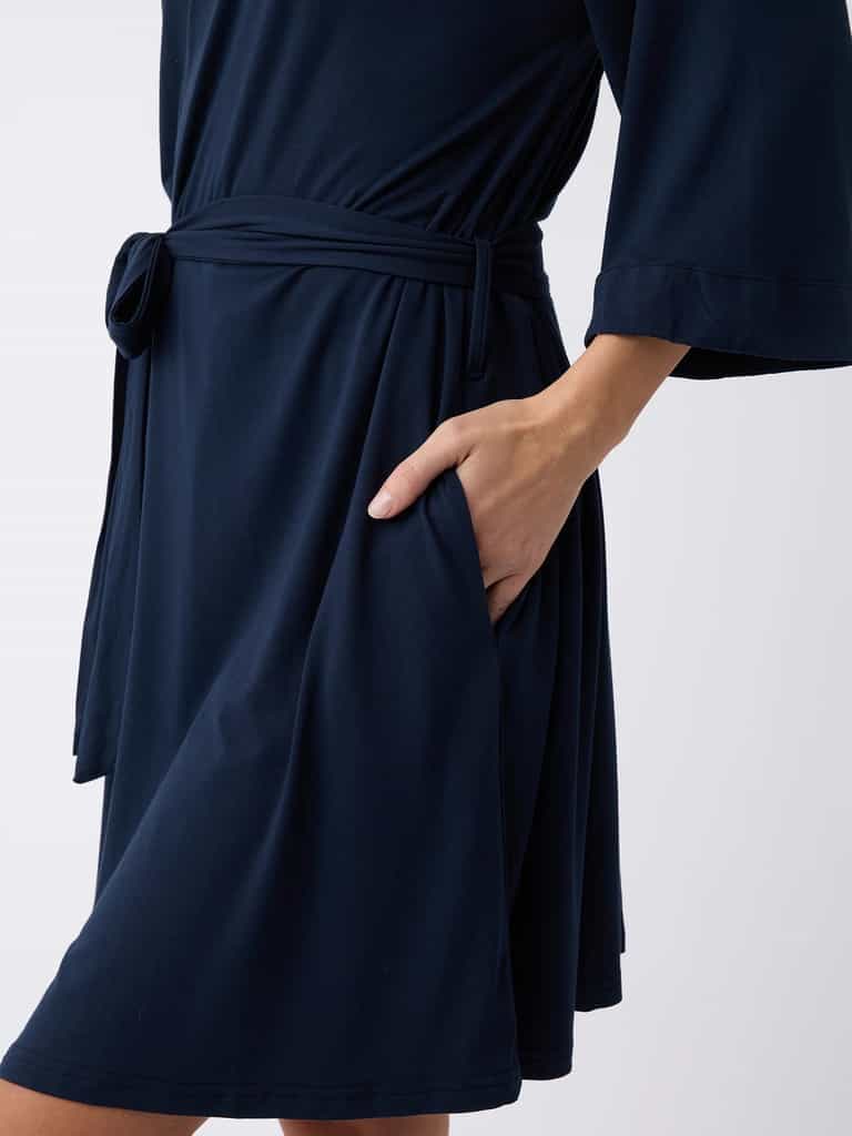 A person is wearing a Women's Stretch-Knit Bamboo Kimono Robe in dark blue from Cozy Earth. The knee-length robe features a waist tie and has the right hand tucked into its side pocket, all set against a plain white background. |Color:Navy
