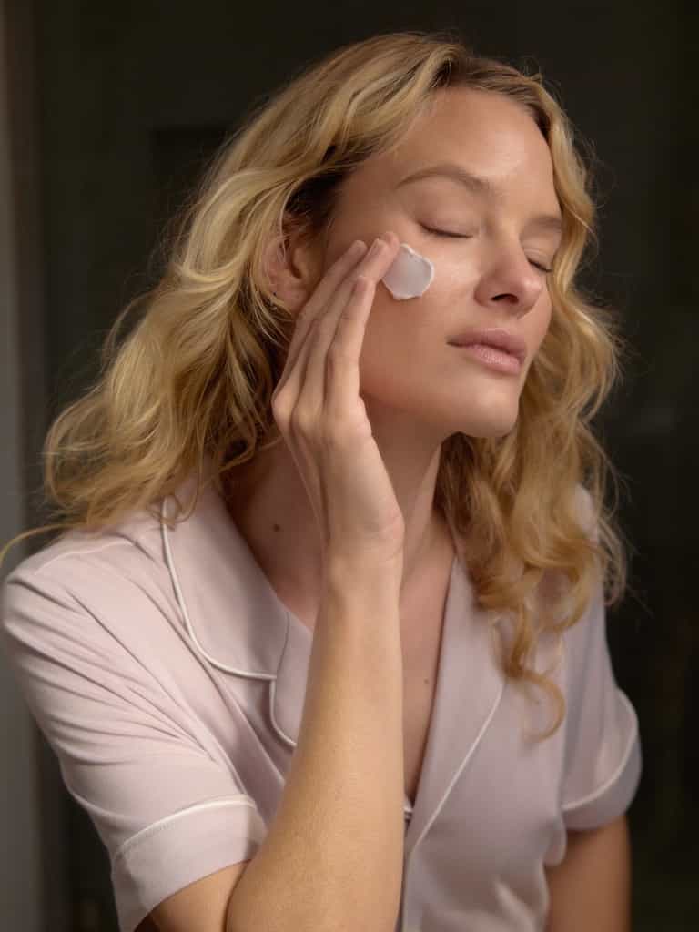 A woman with blonde hair and lilac pajamas spreads a swath of Fount Society Night Cream on her cheek.