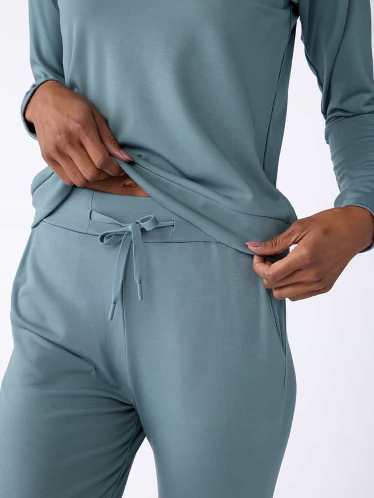 A person wearing the Women's Bamboo Jogger Pant by Cozy Earth, featuring a light blue, long-sleeved top and matching pants with a drawstring waist, gently pulling the shirt. The clothing highlights the soft fabric and relaxed fit. |Color:Oceanside