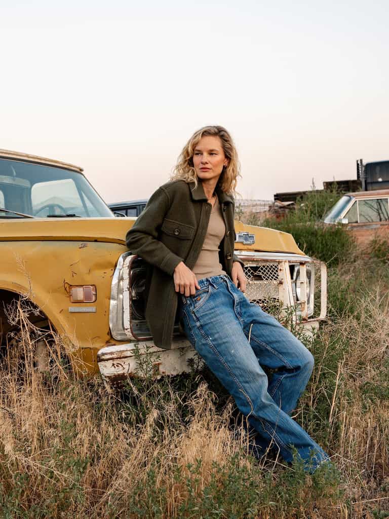 A person with wavy blonde hair leans against a yellow, rusty vintage truck. They are wearing Cozy Earth's Women's Boucle Shacket, a beige top, and blue jeans. The background includes tall grass, an open field, and more old vehicles under a cloudy sky. |Color:Olive