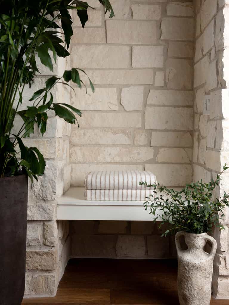 A cozy nook with a white stone wall features a minimalist cream-colored bench topped with two Luxe Bath Towels from Cozy Earth. The space is adorned with two lush green plants, one in a rustic gray pot and the other in an earthenware vase. Natural light highlights the serene setting. |Color:Sand Stripe
