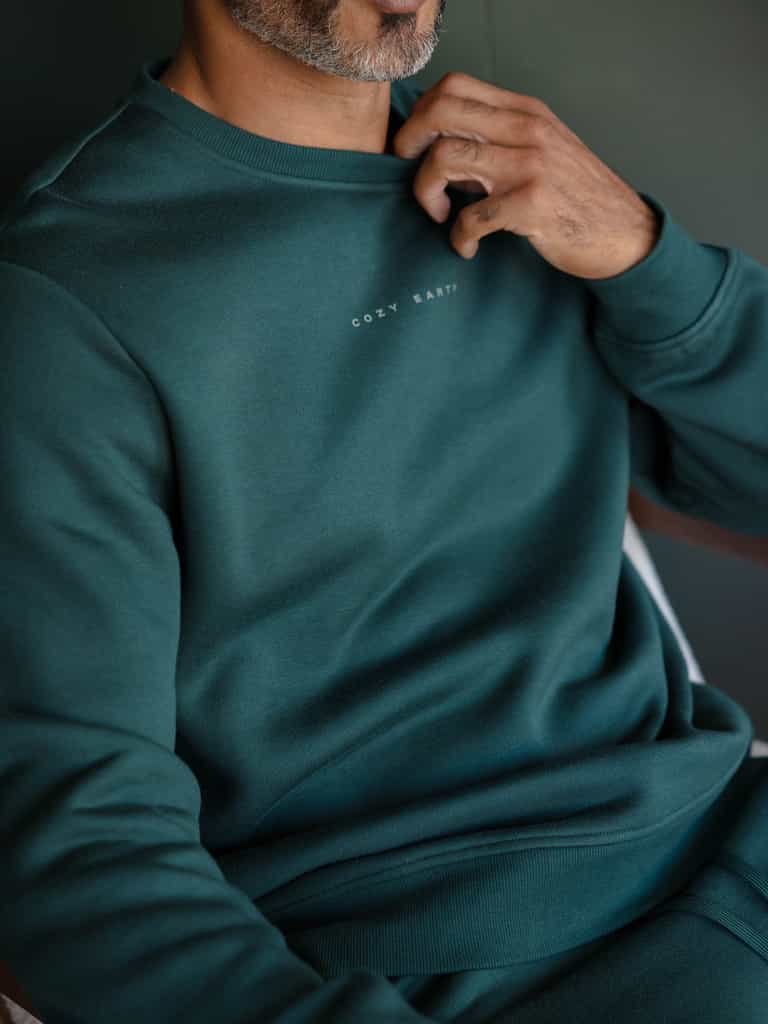A person wearing a dark green Men's CityScape Crewneck with "Cozy Earth" written on the chest, slightly tugging at the neckline. The image highlights the upper body and emphasizes the fabric texture. |Color:Spruce
