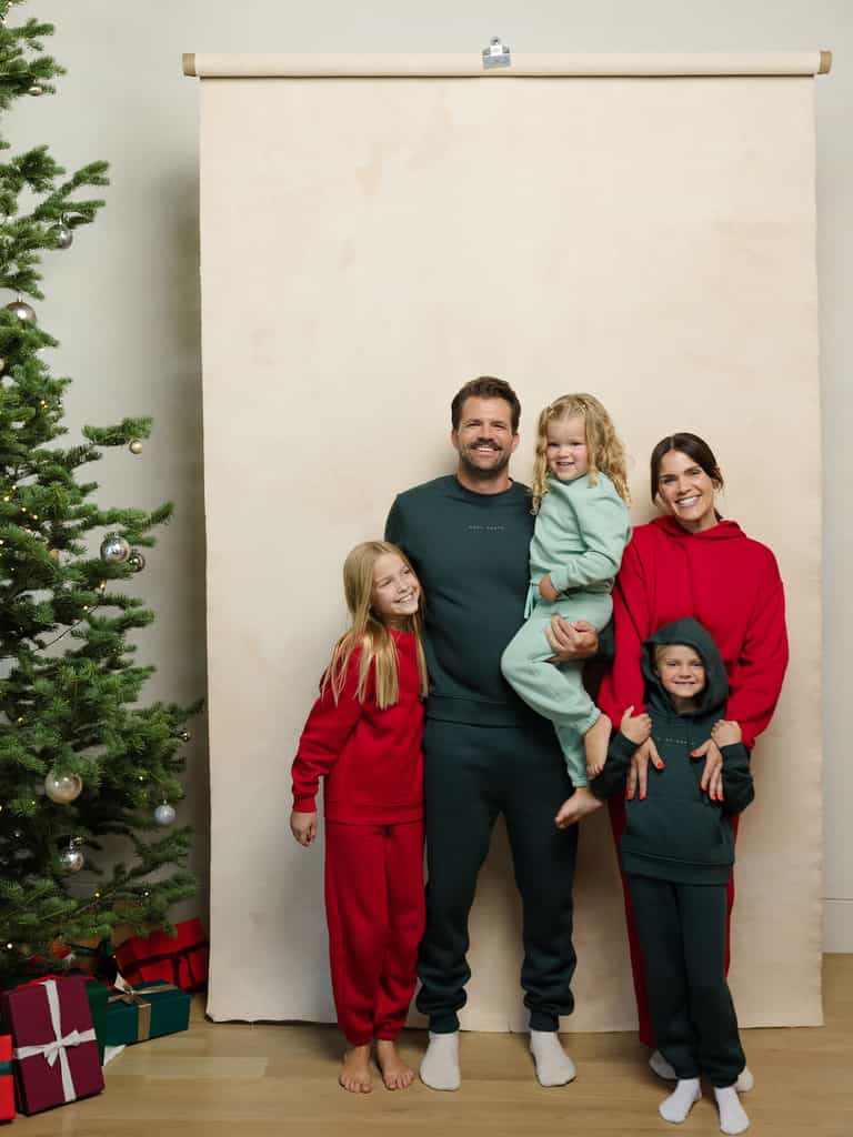 A family of five poses for a holiday photo with the father holding the youngest child. They are all dressed in festive red and green outfits from Cozy Earth. To the left, there's a decorated Christmas tree with presents, set against a beige backdrop. |Color:Spruce