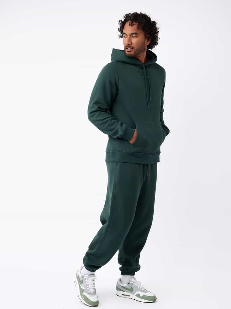 A man stands in a relaxed pose wearing a dark green hoodie paired with Cozy Earth's Men's CityScape Sweatpant. He has one hand in his pocket while looking to the side, and he complements his outfit with white and green sneakers against a white background. |Color:Spruce