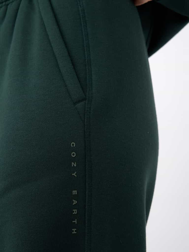 A close-up of a person wearing Women's CityScape Wide Leg Pant in dark green shows the brand name "Cozy Earth" printed vertically along the seam near the pocket. |Color:Spruce
