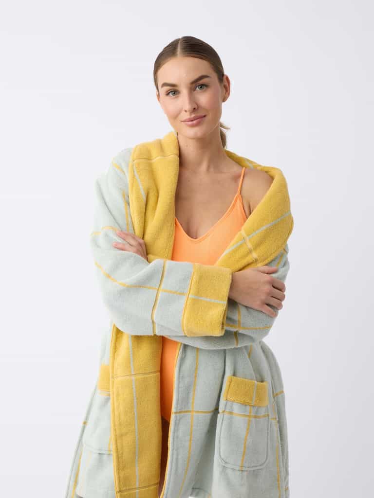 A person with long hair stands against a plain background, smiling with arms crossed while wearing a Cozy Earth Windowpane Resort Robe in light blue with yellow grid patterns and a yellow inner lining over an orange swimsuit. |Color:Sunny Sky