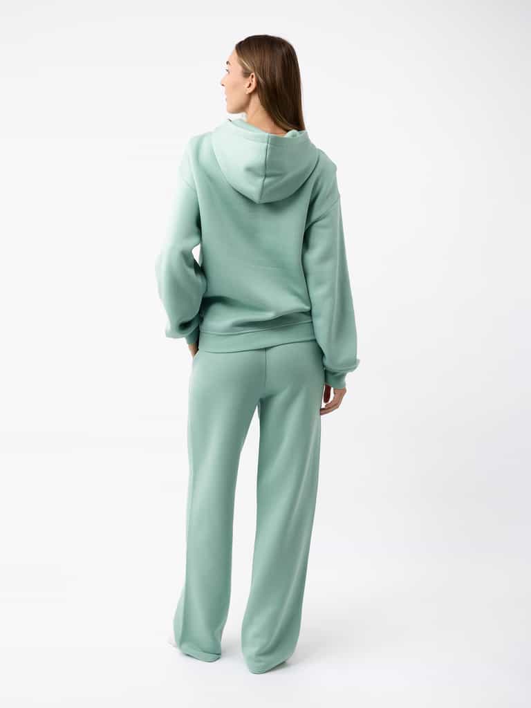 A person with long hair stands facing away, wearing a mint green hoodie and matching Cozy Earth Women's CityScape Wide Leg Pants. The background is a plain white studio setting. The outfit appears casual and comfortable. |Color:Wintergreen