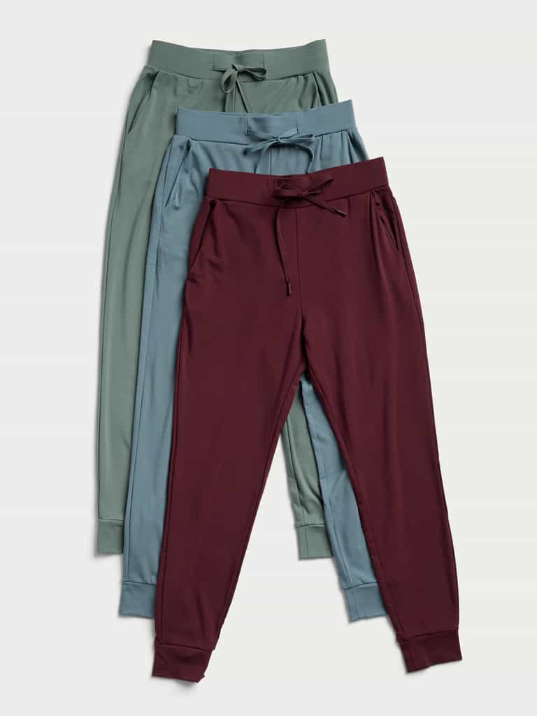 Three pairs of Women's Bamboo Jogger Pants by Cozy Earth in green, teal, and burgundy are laid flat on a light background. Each pair showcases drawstrings and elastic waistbands, complemented by ribbed cuffs at the ankles. The pants are neatly overlapped in a vertical arrangement. |Color:Burgundy