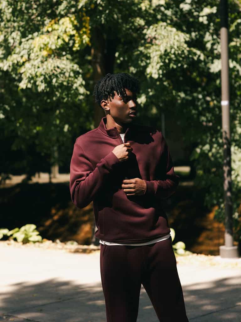 A person is standing outdoors wearing the burgundy Cozy Earth Men's CityScape Quarter Zip with matching pants. They are looking down and touching the collar of their top with one hand. The background includes trees and greenery in a sunlit area. |Color:Burgundy