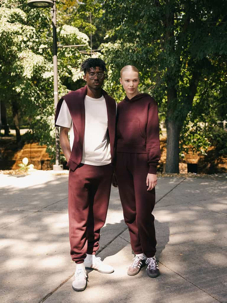 Two individuals stand in an outdoor setting with trees in the background, both wearing Cozy Earth's Men's CityScape Sweatpant in burgundy. The person on the left has a white t-shirt underneath and white sneakers, while the individual on the right sports matching burgundy sneakers. Both look at the camera. |Color:Burgundy