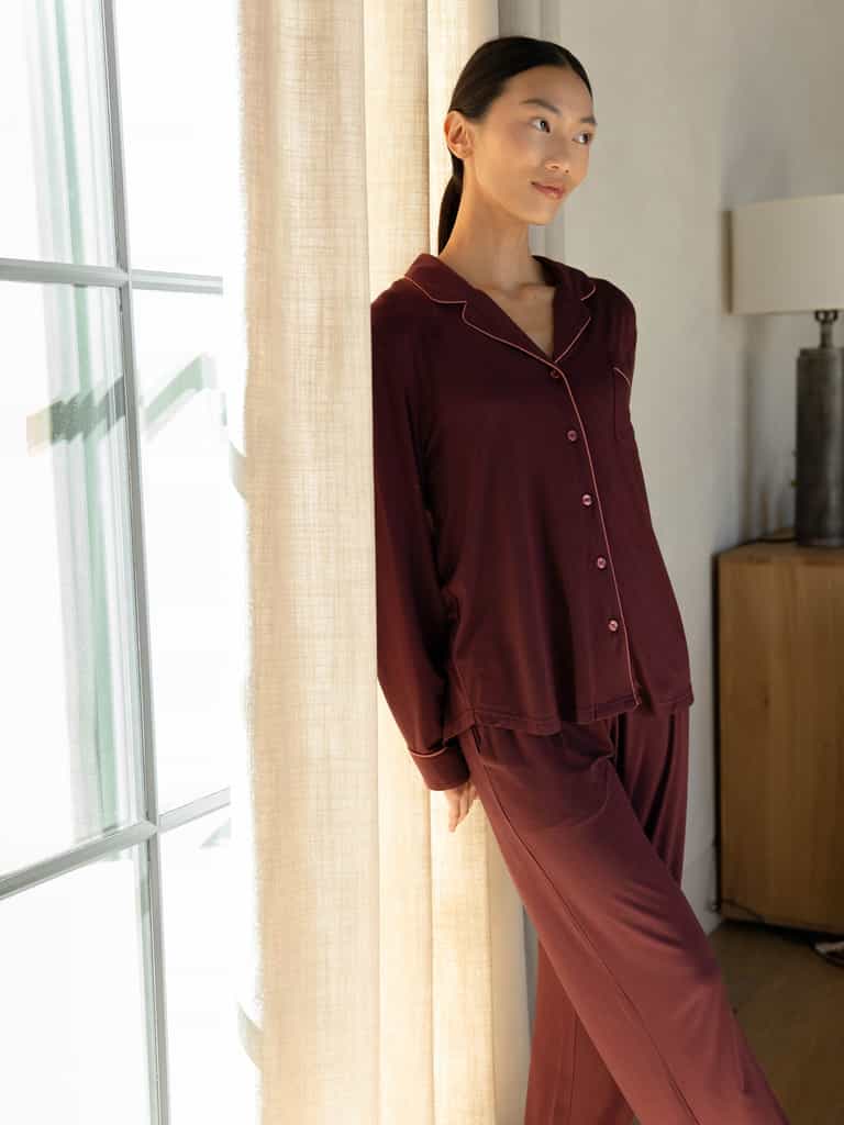 Someone in Cozy Earth's Women's Bamboo Stretch-Knit Long Sleeve Pajama Set, a rich burgundy color, leans thoughtfully against a wall near a curtained window. In the background, there's a lamp and a wooden cabinet. |Color:Burgundy