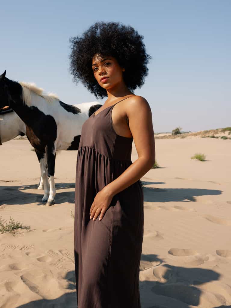 A woman with curly hair wears a loose, dark Cozy Earth Women's Sunset Gathered Dress, standing in a sandy desert landscape. A black and white horse stands in the background under a clear blue sky. |Color:Cacao