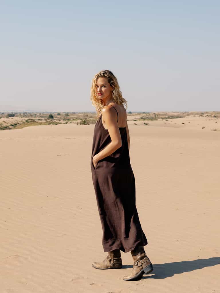 A woman with curly blonde hair, wearing a loose Women’s Sunset Slip Dress from Cozy Earth and brown boots, stands in a sandy desert landscape. She looks over her shoulder at the camera with a slight smile. The sky is clear and blue. |Color:Cacao