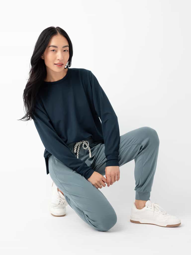A person with long black hair is posing on one knee against a white background. They are wearing a dark blue long-sleeved shirt, Cozy Earth's Women's Studio Jogger in light blue with a drawstring, and white sneakers. |Color:Deep Blue