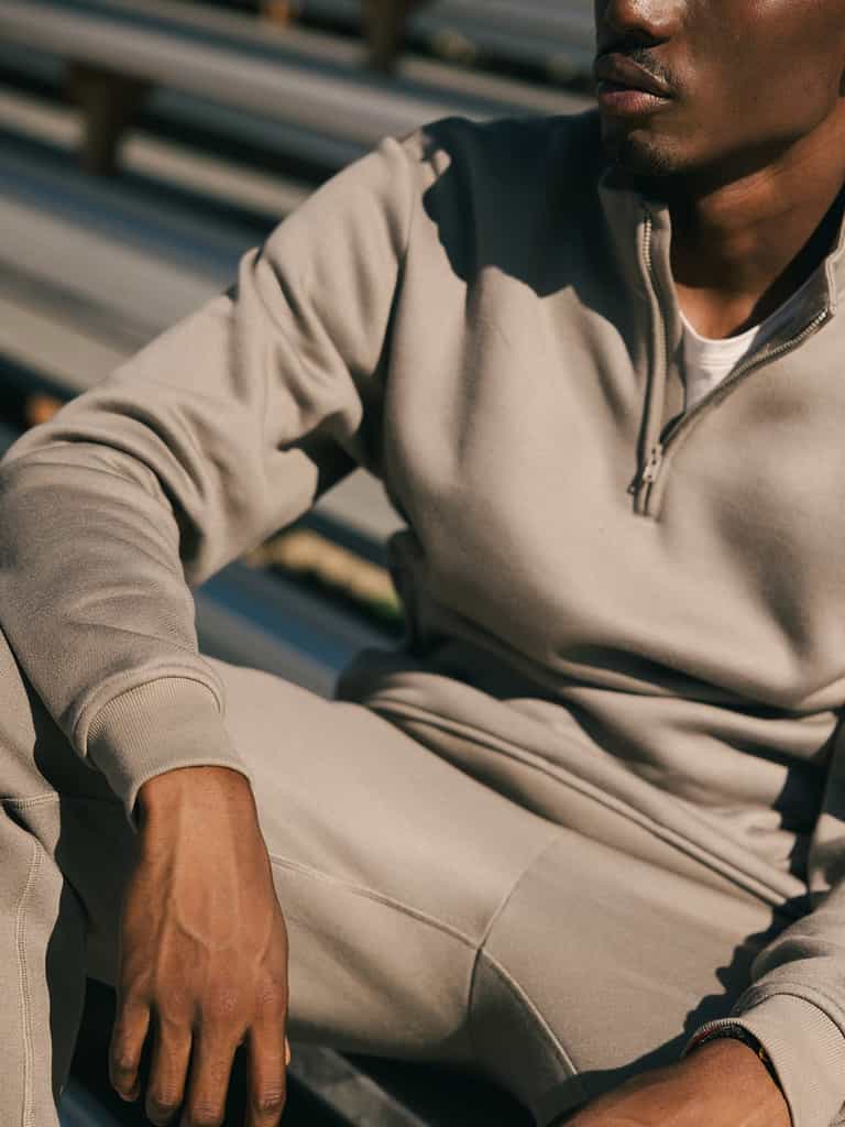A person wearing a Cozy Earth Men's CityScape Quarter Zip in light brown, along with matching pants, is seated outdoors. The image focuses on the upper body and part of the legs, with the person's face partially visible and cropped out of the frame. The background consists of outdoor bleachers. |Color:Ebony