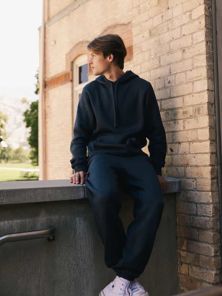 A person wearing a Cozy Earth Men's CityScape Hoodie and sweatpants sits on the edge of a concrete railing, looking to the side. The background features a brick building and trees, with a glimpse of a mountainous landscape. The person is also wearing white sneakers. |Color:Eclipse