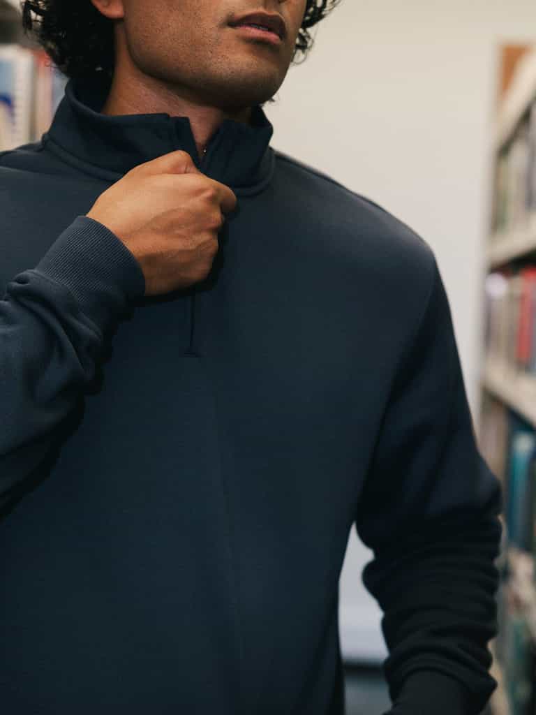 A person with curly hair is depicted from the nose down, wearing a dark blue Men's CityScape Quarter Zip sweater by Cozy Earth. They are adjusting the zipper with one hand. The background features blurred shelves filled with books, suggesting a library or bookstore setting. |Color:Eclipse