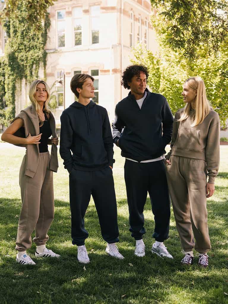 Four people stand conversing on a grassy lawn with trees and a building in the background. They are wearing casual, matching outfits consisting of Cozy Earth's Men's CityScape Sweatpant in earthy tones and white sneakers. The weather appears sunny and pleasant. |Color:Eclipse