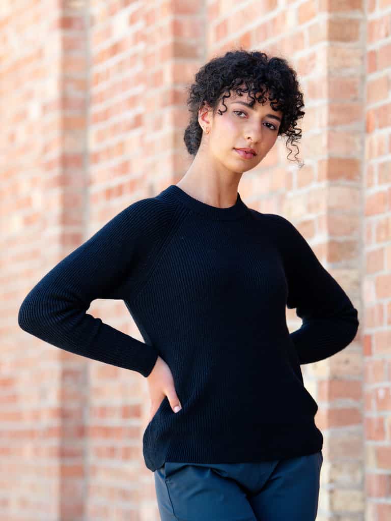 A person with curly hair stands against a brick wall, posing with one hand on their hip and the other by their side. They are wearing a Cozy Earth Women's Classic Crewneck and dark pants. The expression on their face is neutral. |Color:Jet Black