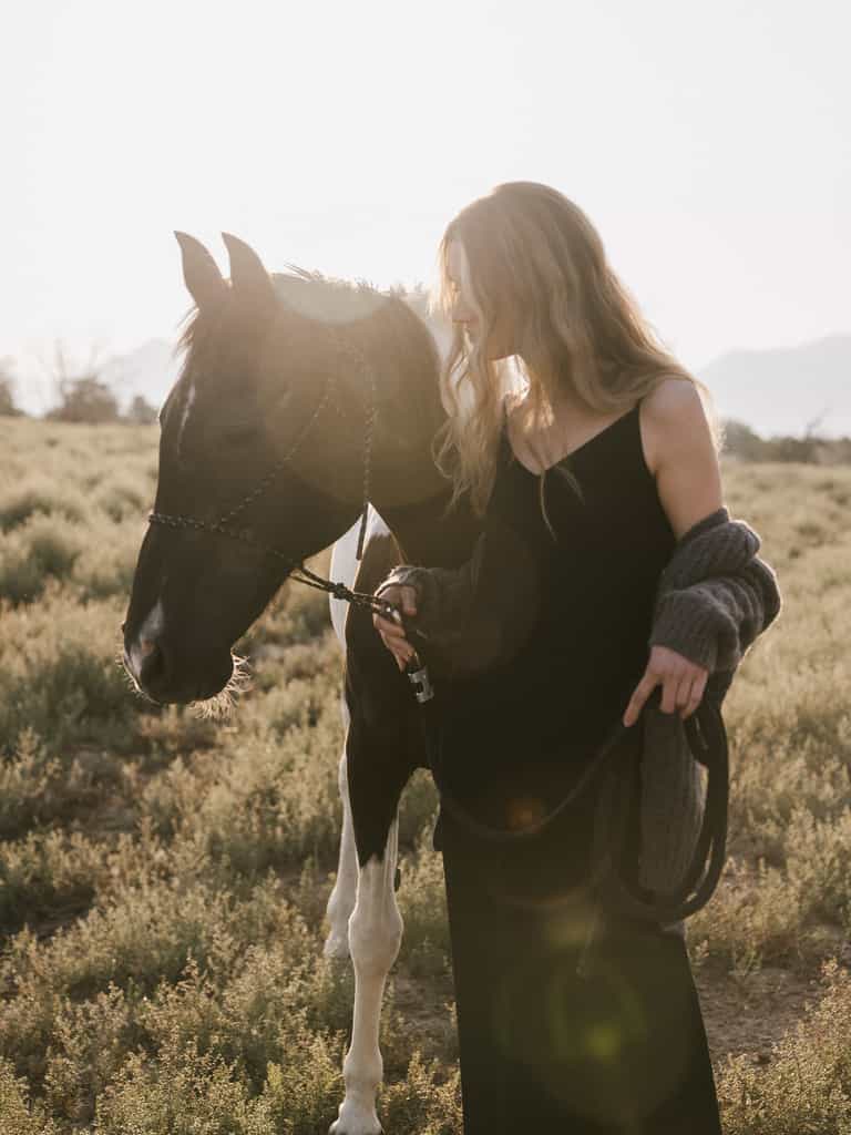 A woman wearing the Women's Sunset Slip Dress by Cozy Earth and a gray sweater stands in a grassy field at sunset, gently holding the reins of a black and white horse. With their heads slightly bowed, sunlight filters through the scene, casting a warm glow. |Color:Jet Black