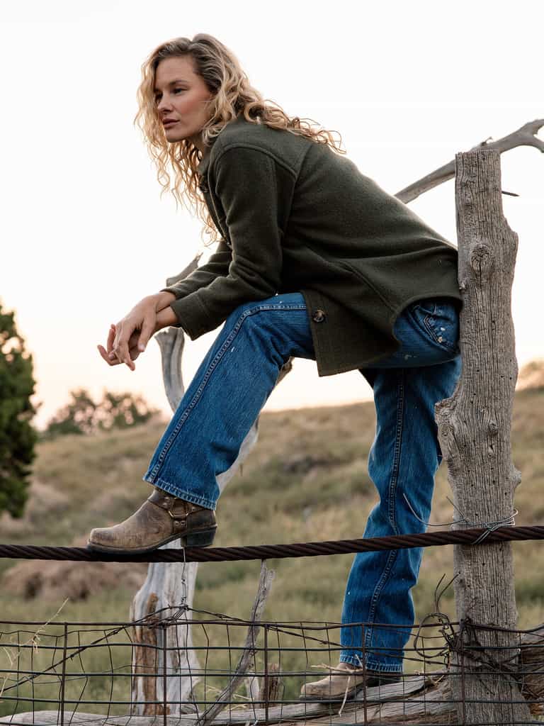 A person with long curly hair, wearing a Cozy Earth Women's Boucle Shacket, blue jeans, and brown boots, is sitting on a rustic wooden fence in a rural outdoor setting. The person appears to be gazing into the distance as the sun sets. |Color:Olive